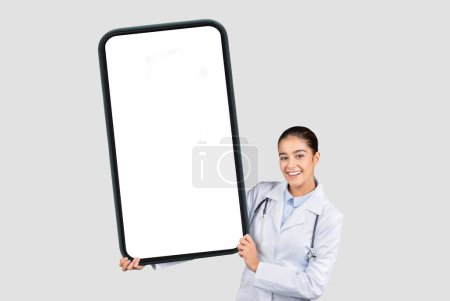 Photo for Cheerful caucasian young woman doctor in white coat show big smartphone with blank screen, isolated on gray studio background, collage. Medical health care app, website recommendation - Royalty Free Image