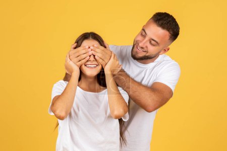 Photo for Happy young european man in white t-shirt, closes eyes to woman for surprise, isolated on yellow studio background. Romantic relationships and love, birthday, anniversary present - Royalty Free Image