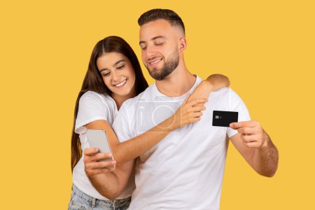 Photo for Glad millennial european lady shopaholic hug man in white t-shirts, with credit card and phone, isolated on yellow studio background. Recommendation shopping online app, cashback - Royalty Free Image
