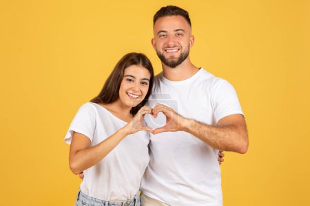 Photo for Cheerful young european man and woman in white t-shirts hugs, making heart gesture with hands, enjoy free time and love, isolated on yellow studio background. Emotions date and romantic lifestyle - Royalty Free Image