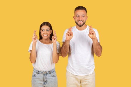Photo for Happy millennial european wife and husband in white t-shirts with crossed fingers hoping or wishing, isolated on yellow studio background. Optimistic lifestyle, dream come true - Royalty Free Image