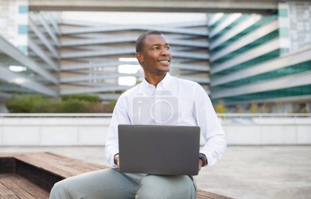 Téléchargez les photos : Homme d'affaires noir souriant utilisant un ordinateur portable tout en étant assis sur un banc extérieur sur fond de bâtiment moderne, entrepreneur homme afro-américain rêveur tenant l'ordinateur et détournant les yeux, copier l'espace - en image libre de droit