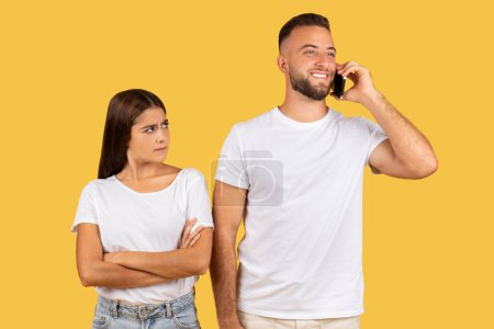 Photo for Angry young european lady in white t-shirt, look at happy man, calls by phone, isolated on yellow studio background. Relationships problems, communication, betrayal and jealousy - Royalty Free Image