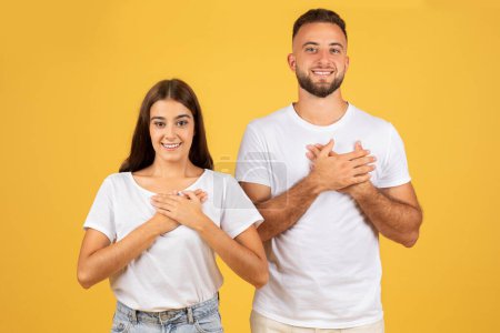 Photo for Glad young european man and woman in white t-shirts put hands to chest to heart, enjoy love, isolated on yellow studio background. Emotions gratitude, thanks and romantic lifestyle - Royalty Free Image