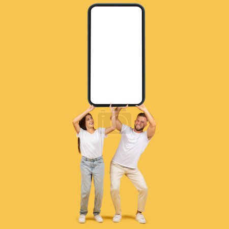 Photo for Happy funny millennial european family in white t-shirts hold huge smartphone with empty screen, isolated on yellow studio background. Online shopping, app, sale website, ad and offer - Royalty Free Image