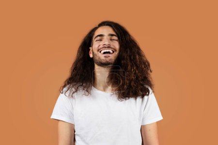 Foto de Joven hispano alegre con el pelo castaño rizado se ríe y sonríe en pose casual, de pie contra el fondo del estudio beige, usando una camiseta blanca. Chico expresando emociones positivas y felicidad - Imagen libre de derechos