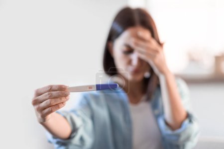 Photo for Upset young lady holding positive pregnancy test at home, covering face in distress and worry over unexpected motherhood, reflecting on difficult decision ahead. Selective focus - Royalty Free Image