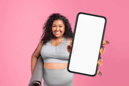 Foto de Aplicación de entrenamiento publicitario de dama negra de gran tamaño, que muestra un gran teléfono celular con pantalla vacía, sujetando la estera del gimnasio sobre el fondo del estudio rosa. Tecnología en pérdida de peso y fitness - Imagen libre de derechos
