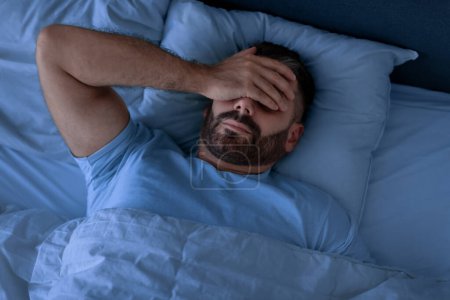 Photo for Insomnia Problem. Man Covering Face With Hand Lying Unable To Sleep In Bed At Home, Suffering From Headache And Sleeplessness In Modern Bedroom Interior. Top View, Low Light - Royalty Free Image