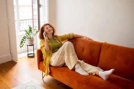 Foto de Mujer mayor pelirroja feliz hablando en el teléfono celular mientras se relaja en el sofá en casa, sonriendo anciana descansando en el acogedor interior de la sala de estar, disfrutando de la agradable conversación del teléfono móvil, Copiar el espacio - Imagen libre de derechos
