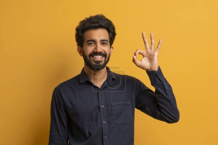Photo for Young smiling indian man making OK gesture at camera, joyful handsome eastern guy gesturing okay, cheerful male showing sign of approval while standing against yellow background, copy space - Royalty Free Image
