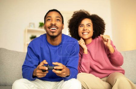 Téléchargez les photos : Jeune couple afro-américain profitant d'une séance de jeu amusante avec une manette numérique, jouant au jeu vidéo ensemble pendant le week-end, se détendre sur leur canapé à l'intérieur de la maison. Technologie et divertissement - en image libre de droit