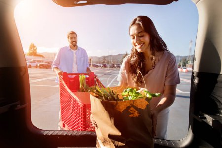 Téléchargez les photos : Jeune femme arabe déballe joyeusement épicerie fraîche du sac en papier dans son coffre de voitures que l'homme heureux approche avec un chariot sur fond, vue de l'intérieur automatique - en image libre de droit