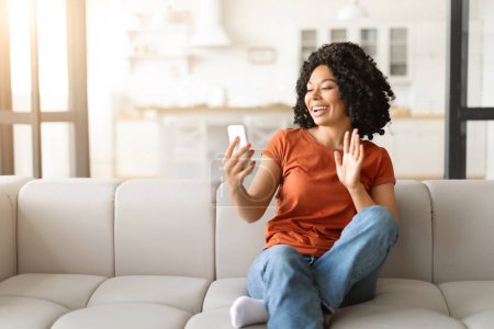 Téléchargez les photos : Joyeux jeune femme noire faisant appel vidéo sur Smartphone à la maison, afro-américaine dame agitant la main à la caméra de téléphone portable tout en se relaxant sur le canapé, profiter de la communication en ligne, espace libre - en image libre de droit