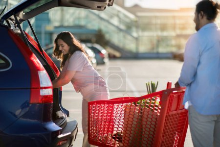 Téléchargez les photos : Jeune femme plaçant des produits d'épicerie dans le coffre de la voiture tandis que l'homme se tient prêt avec un panier complet dans un parking ensoleillé à l'extérieur après des achats réussis - en image libre de droit