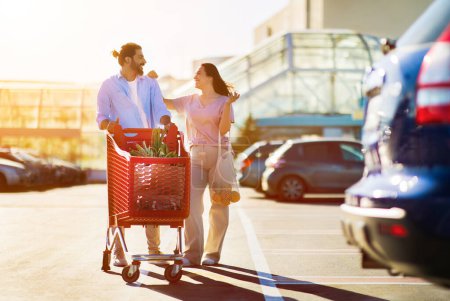 Photo for Happy arab man and latin woman with full grocery cart walking together in a sunny parking lot, having a cheerful conversation, full length - Royalty Free Image