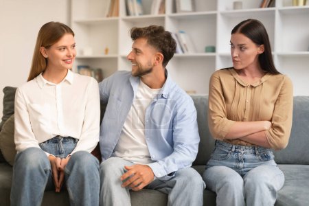 Photo for Young man hugs his girlfriend on couch while another woman looks with jealousy crossing hands, sitting together on sofa in modern living room. Infidelity, complicated romantic triangle - Royalty Free Image