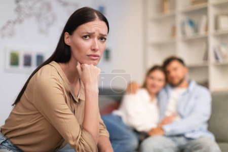 Photo for Millennial couple embraces on couch while unhappy young lady looking at camera, suffering from loneliness and jealousy, complexity of friendship relationships between lonely and married friends - Royalty Free Image