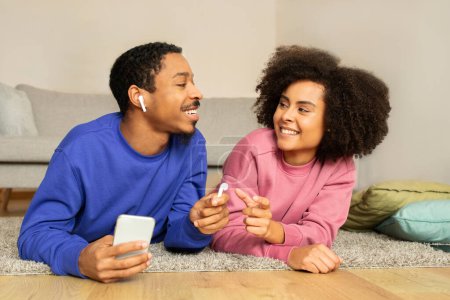Téléchargez les photos : Jeune couple afro-américain se détend ensemble écouter de la musique sur le téléphone en utilisant des écouteurs, profiter de loisirs de détente domestique et mode de vie numérique, couché sur le sol Dans leur intérieur confortable salon - en image libre de droit
