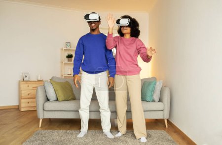 Foto de Pareja negra joven totalmente inmersa en el juego de realidad virtual en casa, con auriculares VR y juguetoneando con entretenimiento digital innovador en su sala de estar. Futuro de la tecnología de juego - Imagen libre de derechos