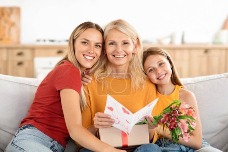 Photo for An affectionate grown daughter and her junior granddaughter warmly greet their granny on Mothers Day, delighting her with a gift, fresh flowers, and a personally crafted card - Royalty Free Image