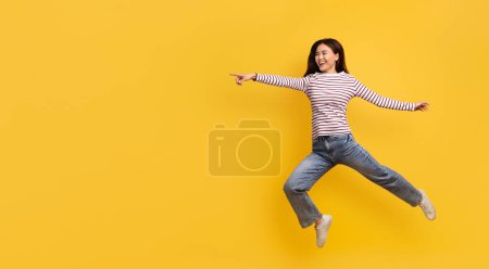 Foto de Divertida joven coreana saltando en el aire y apuntando al espacio de copia para la publicidad aislada en el fondo del estudio amarillo, mostrando una gran oferta, buen trato, panorama, longitud completa - Imagen libre de derechos