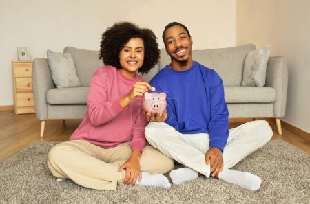 Téléchargez les photos : Joyeux conjoints noirs tenant et mettant de l'argent dans la tirelire assis dans leur salon intérieur, souriant à la caméra. Objectifs financiers familiaux et concept de stratégie d'épargne - en image libre de droit