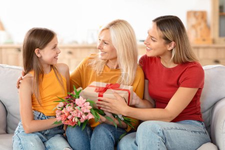 Téléchargez les photos : Joyeuse famille caucasienne de trois générations partageant un moment de fête des mères à la maison. Grand-mère souriante tient des fleurs et un cadeau de sa fille et petite-fille - en image libre de droit