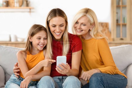 Téléchargez les photos : Famille de trois générations profitant du temps avec un smartphone. Grand-mère, mère et fille souriant en regardant un téléphone portable, partageant un moment de liaison ensemble - en image libre de droit