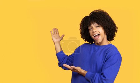 Téléchargez les photos : Joyeux jeune homme noir pointant vers l'espace de copie avec les paumes ouvertes, heureux homme afro-américain présentant quelque chose, démontrant place libre pour votre publicité, debout sur fond jaune - en image libre de droit