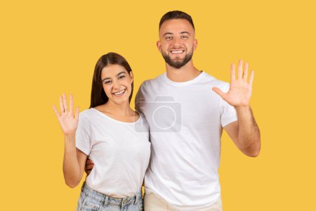 Photo for Happy millennial european wife and husband bloggers in white t-shirts waving hands, say hello, isolated on yellow studio background. Communication gesture, video call, ad and offer - Royalty Free Image