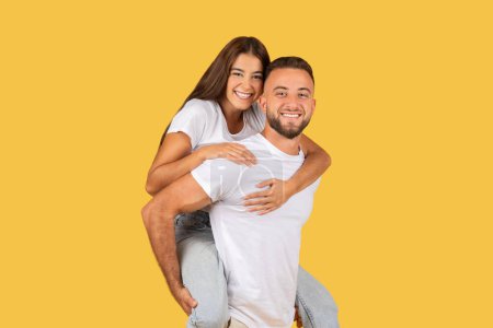 Photo for Cheerful millennial european guy in white t-shirt hold lady on back, play together, enjoy free time, have fun, isolated on yellow studio background. Lifestyle, relationships and love - Royalty Free Image