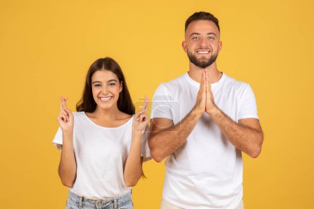 Photo for Smiling young european man and woman in white t-shirts with crossed fingers hoping or wishing and make pray gesture, isolated on yellow studio background. Emotions and lifestyle, dream come true - Royalty Free Image