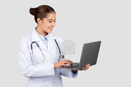 Photo for Positive millennial european lady doctor typing on computer, chatting with patient, in white coat on light background. Medical services app, exam, professional health care and help with gadget - Royalty Free Image