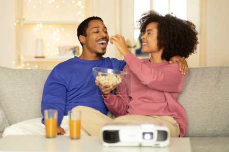 Téléchargez les photos : Jeune couple noir profite d'un week-end amusant à la maison, regarder un film sur leur projecteur, femme nourrit mari pop-corn, alors qu'ils rient et se détendre sur le canapé dans l'intérieur du salon moderne - en image libre de droit
