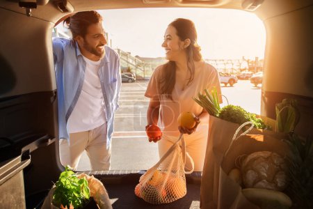 Téléchargez les photos : Jeune couple moyen-oriental partage un moment de joie tout en inspectant les poivrons frais et les oranges par le coffre de leur voiture dans le parking - en image libre de droit