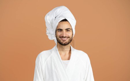 Téléchargez les photos : Portrait d'un homme hispanique souriant en peignoir blanc avec serviette enveloppée sur la tête sur fond brun, jeune homme posant en studio après la douche du matin et routine d'auto-soins - en image libre de droit