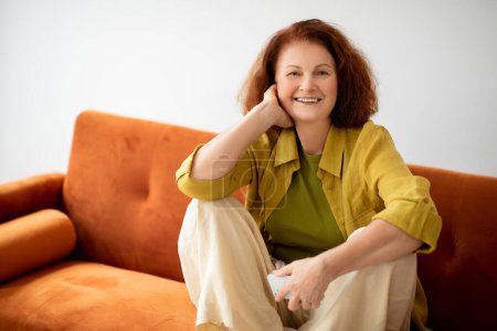 Téléchargez les photos : Belle femme âgée avec Smartphone dans la main se détendre sur un canapé confortable à la maison, Happy Redhead âgée femelle se reposant dans le salon intérieur, profiter du temps de retraite, sourire à la caméra - en image libre de droit