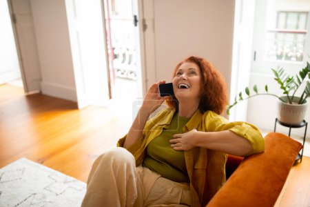 Foto de Feliz jubilación. Mujer mayor despreocupada riendo mientras habla en el teléfono celular en casa, señora anciana positiva disfrutando de la conversación del teléfono móvil con un amigo mientras se relaja en el sofá en la sala de estar - Imagen libre de derechos