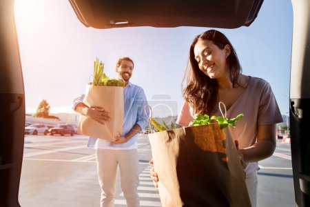 Téléchargez les photos : Homme arabe souriant avec des sacs d'épicerie à la main que la jeune femme regarde joyeusement verts frais, les mettant dans le coffre de la voiture, vue de l'intérieur de l'automobile - en image libre de droit