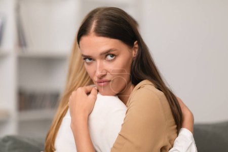 Photo for Annoyed young lady embracing her girlfriend with displeased expression indoor, fake friendship concept. Two women friends hugging, crying lady looking for support from angry indifferent pal - Royalty Free Image