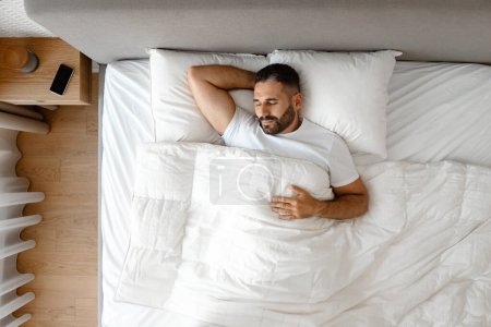 Photo for Above view of peaceful middle aged man enjoying nap in his modern bedroom. Guy lying comfortably in bed holding hand behind head, wrapped in cozy linens. Rest and relaxation, healthy sleep - Royalty Free Image