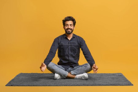 Foto de Hombre indio joven relajado con ropa casual meditando solo, sentado con las piernas cruzadas en la alfombra, chico oriental guapo practicando yoga, posando aislado sobre fondo de estudio amarillo, espacio de copia - Imagen libre de derechos
