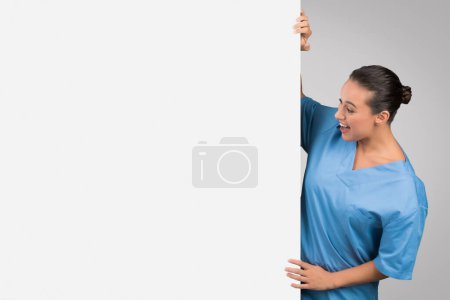 Foto de Trabajadora médica joven mirando el tablero blanco enorme de la publicidad con el espacio libre para el texto o el diseño, de pie sobre fondo claro, bandera - Imagen libre de derechos