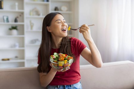 Foto de Mujer asiática feliz disfrutando de ensalada de verduras frescas en casa, alegre joven coreana sosteniendo un tazón con comida vegetariana saludable, comiendo sabroso almuerzo mientras está sentado en el sofá en la sala de estar, espacio para copiar - Imagen libre de derechos