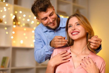 Téléchargez les photos : Une femme joyeuse reçoit un délicat collier de cœur d'un homme aimant, capturant un moment cadeau surprise dans une pièce chaude et éclairée avec des guirlandes, espace libre - en image libre de droit