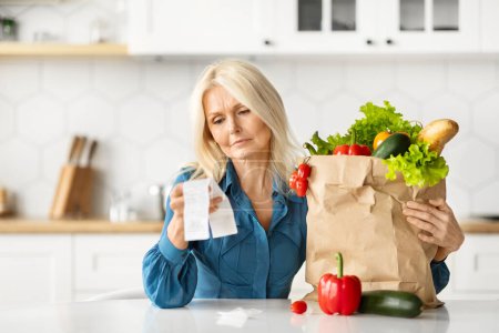 Foto de Confundido Senior Housewife Checking Grocery Bills In Kitchen After Food Shopping, Mujer anciana molesta preocupada por los precios caros de verduras y frutas orgánicas, Primer plano Shot - Imagen libre de derechos