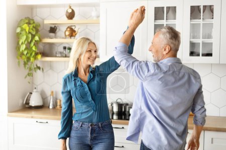 Téléchargez les photos : Portrait de joyeux couple âgé dansant ensemble à l'intérieur de la cuisine, heureux époux aînés romantiques s'amusant, se tenant la main et se souriant, appréciant passer du temps à la maison - en image libre de droit