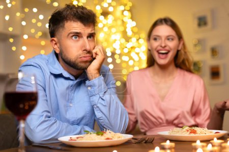Foto de Hombre que parece aburrido descansando su mejilla en la mano en una mesa de cena, mientras que la mujer a su lado se ríe, creando un ambiente contrastante con luces festivas en el fondo - Imagen libre de derechos