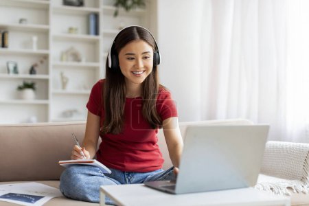 Téléchargez les photos : Enseignement à distance. Jeune femme asiatique souriante dans les écouteurs sans fil étudiant avec ordinateur portable à la maison, heureuse coréenne utilisant l'ordinateur pour l'éducation en ligne tout en étant assis sur le canapé dans le salon - en image libre de droit